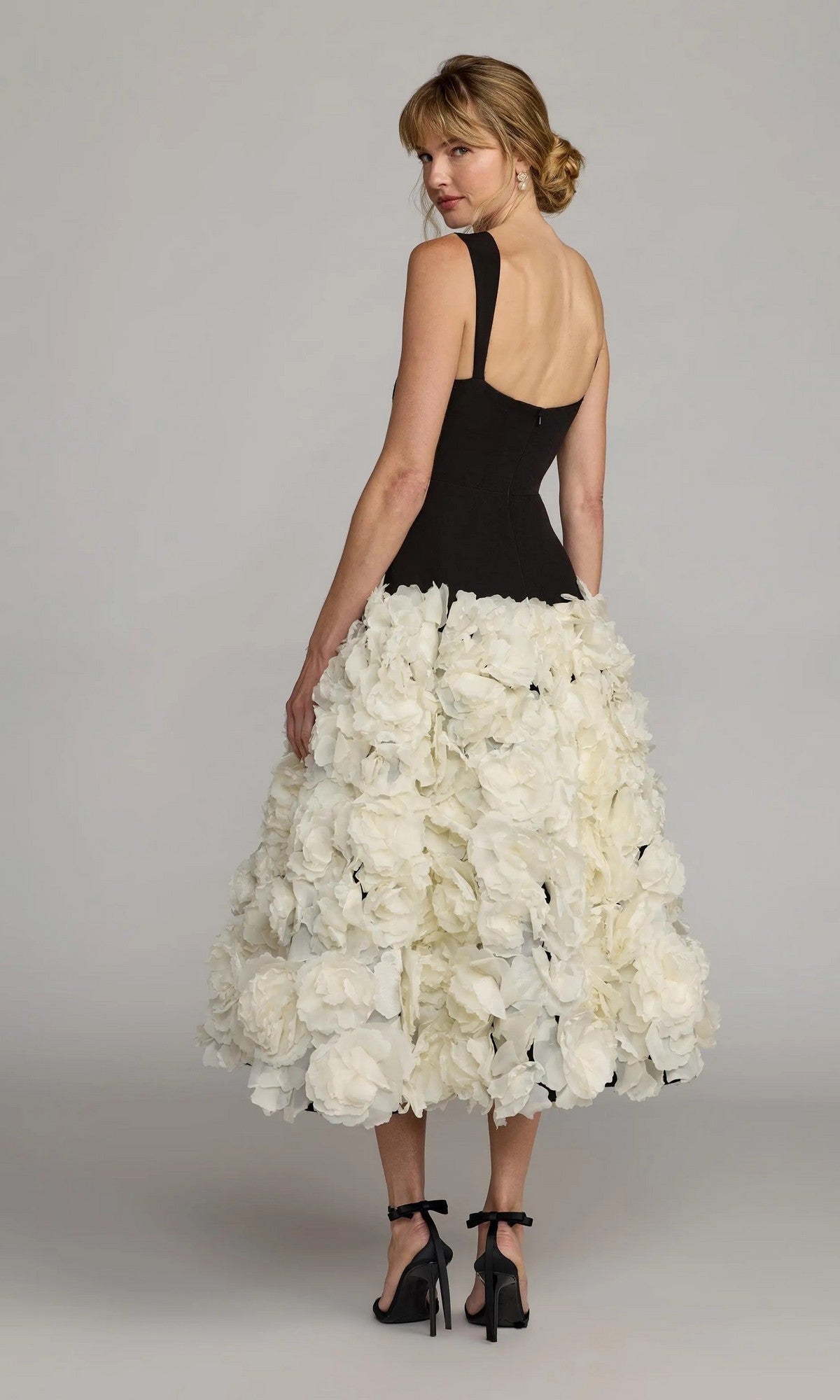 A woman stands in front of a plain backdrop, wearing the Mac Duggal 21080 Midi Wedding Guest Dress—a sleeveless black crepe design with a voluminous skirt embellished with 3D organza flowers—glancing over her shoulder.